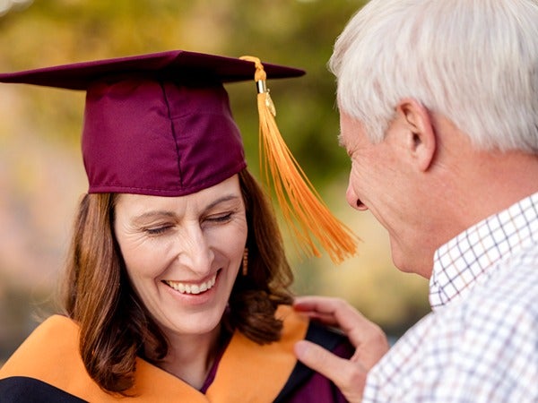 A woman in a maroon graduation cap and gown smiles next to an older man outdoors.