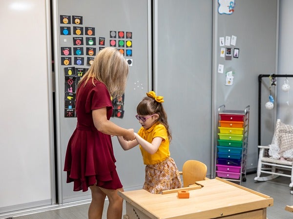 A woman interacts with a young girl in a classroom setting, with colorful symbols and furniture around them.