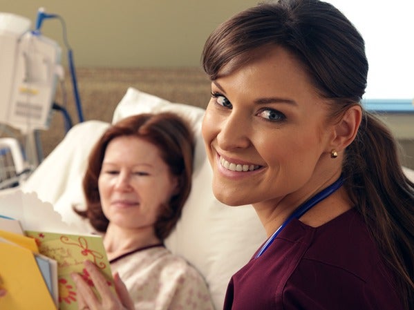 A medical professional smiling at the camera beside a patient in a hospital bed reading cards.