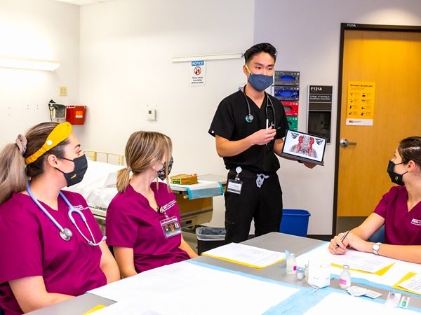 Four medical students in scrubs discussing around a table; one is holding a tablet with a medical diagram.
