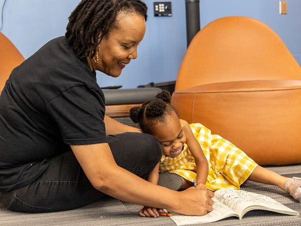 A woman and a girl coloring together on the floor in a cozy room with bean bag chairs.
