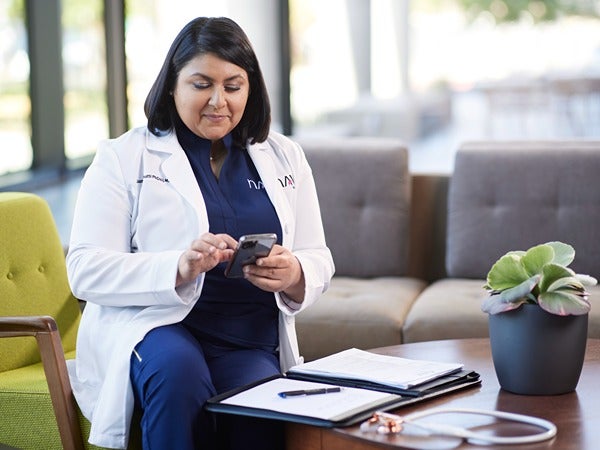 A person in a lab coat using a smartphone, seated near a table with a notebook, stethoscope, and a plant.