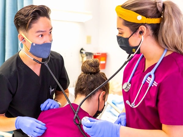 Three people in a medical setting, two wearing maroon scrubs and using stethoscopes.