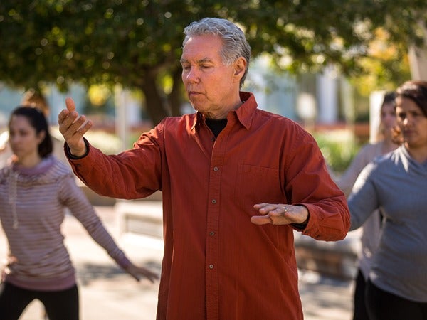 A man in a red shirt performs tai chi outdoors with others.