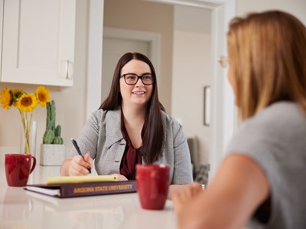 Two people conversing at a kitchen counter, with a book labeled "Arizona State University" visible.