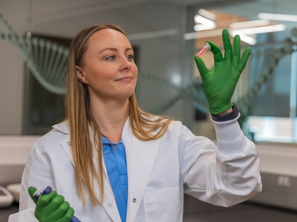 A woman in a lab coat and green gloves examines a test tube in a laboratory.