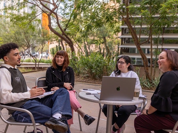 Four people sit at an outdoor table having a discussion, with a laptop and papers on the table.