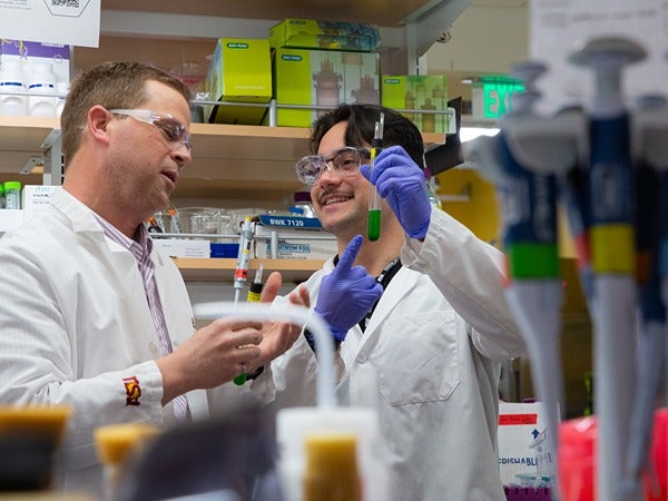 Two people in a laboratory wearing lab coats and goggles, holding a pipette and a test tube.