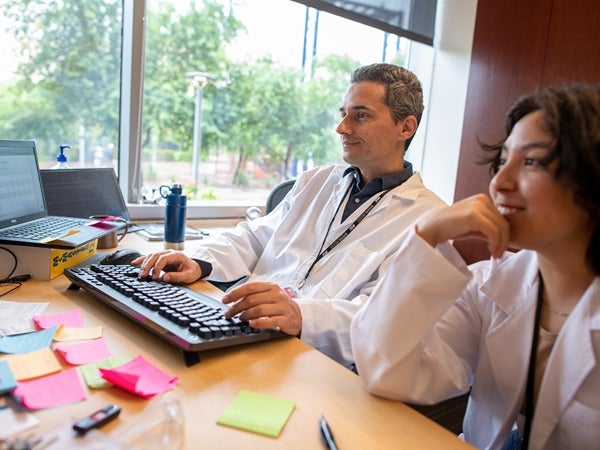 Two people in lab coats working at a computer in an office with a window in the background.