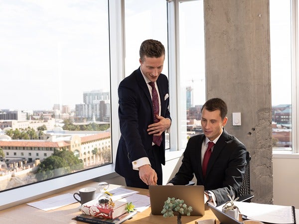 Two men in business suits in an office with a city view, discussing something on a laptop.