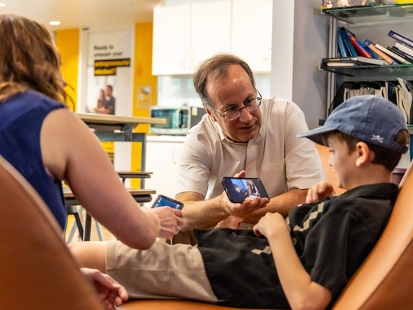 A man shows a smartphone to a young boy in a chair, with a woman seated nearby.