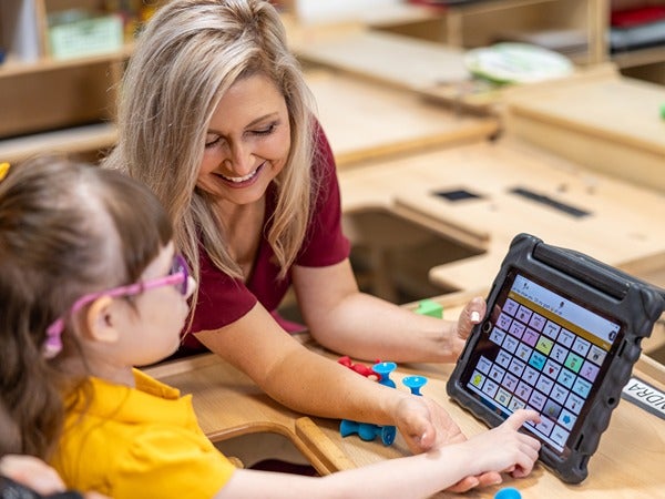 A child with glasses and a woman are using a touchscreen tablet in a classroom.
