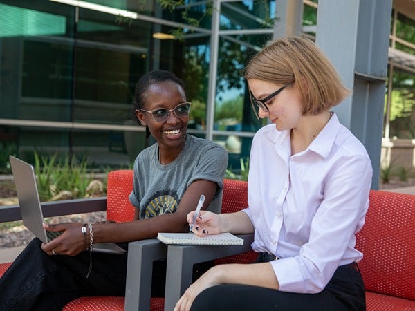 Two people sitting outside, interacting with a laptop and a notebook.