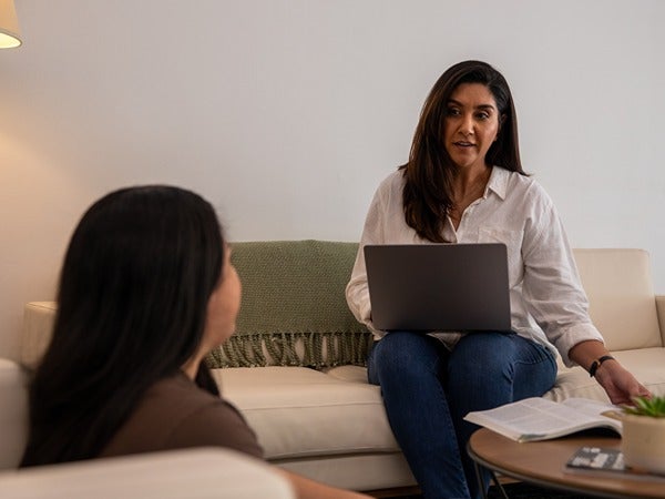 Two women in a living room, one with a laptop, conversing on a sofa.