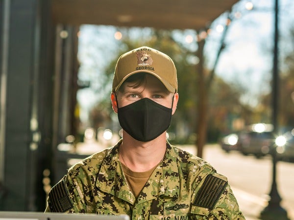 Person in military camouflage uniform wearing a black face mask and beige cap outdoors.