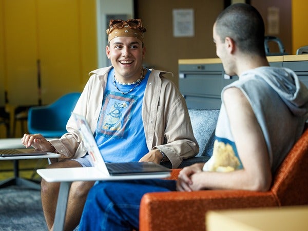 Two people sitting and talking in a lounge area with laptops.
