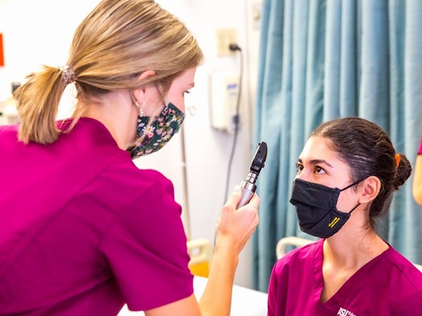 Two individuals in maroon scrubs, one examining the other's eyes with an ophthalmoscope in a clinical setting.