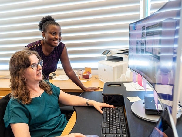 Two women in an office, one seated at a computer, the other standing beside her.