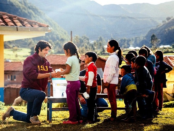 A man gives a package to a girl in a rural setting, surrounded by a line of children.