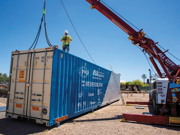 A crane lifts a blue shipping container with a worker standing on top.