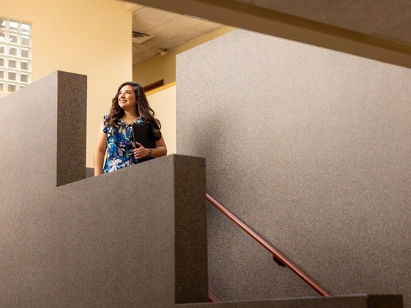A woman in a blue floral blouse stands on a staircase, looking up and smiling, holding a folder.