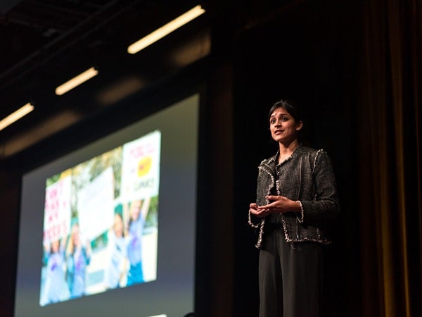 Person presenting on a stage with a projected image in the background.