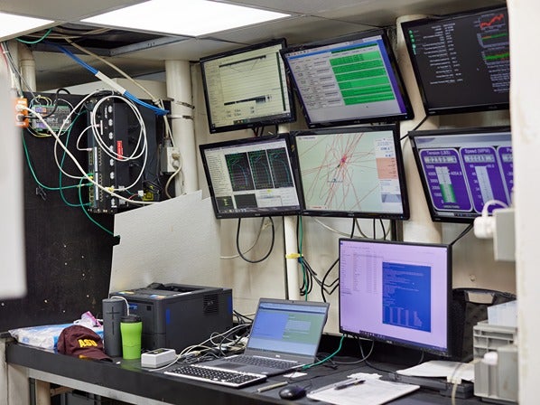 A technical monitoring room with multiple screens displaying data and graphs, a desk with electronic devices, cables, a laptop, and a printer.