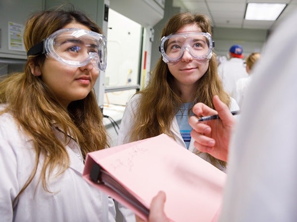 Two women in lab coats and goggles in a laboratory setting.