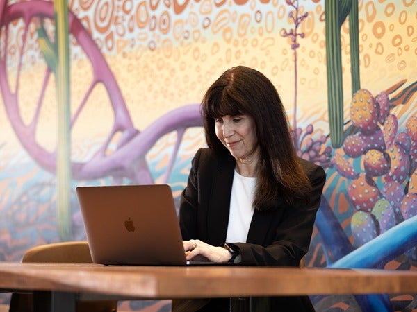 A woman in a black blazer works on a laptop in front of a colorful mural.