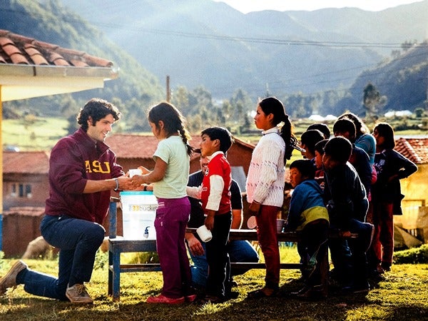 Person in maroon hoodie kneeling, interacting with children in a rural village setting with mountains in the background.