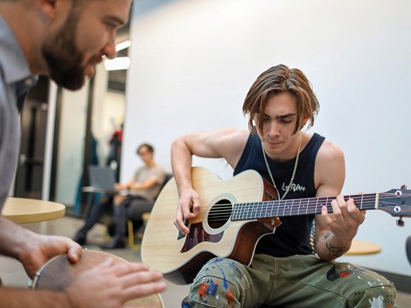 Young man playing an acoustic guitar while another person plays a hand drum nearby.