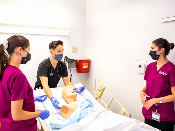 Three people in medical scrubs and masks practice with a mannequin on a hospital bed.