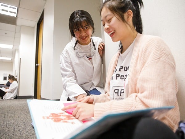 Two women sitting on the floor, discussing a book with an anatomy illustration in a hallway.