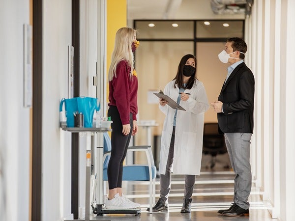 Three people conversing in a hallway, with one standing on a scale and another taking notes.