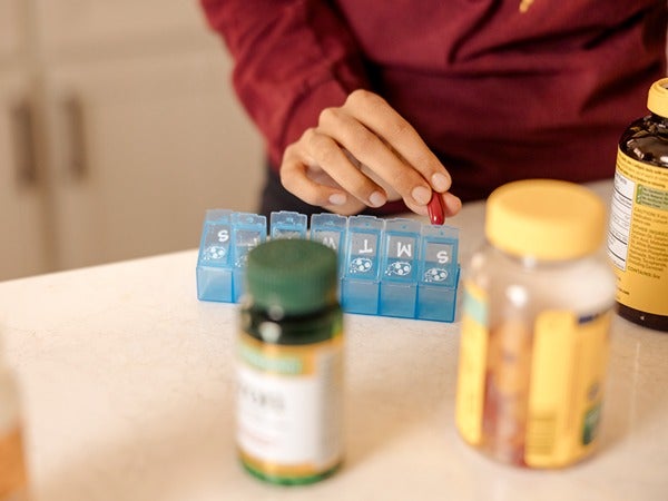 A person placing a red pill in a blue pill organizer with pill bottles nearby.
