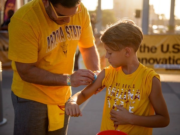 A man in a gold "Arizona State" shirt swabs a boy's arm outdoors.