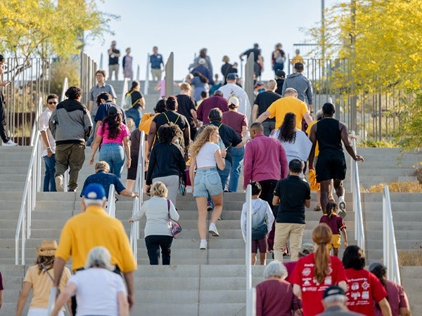 A crowd of people walking up a large staircase outdoors on a sunny day, with trees and clear sky in the background.