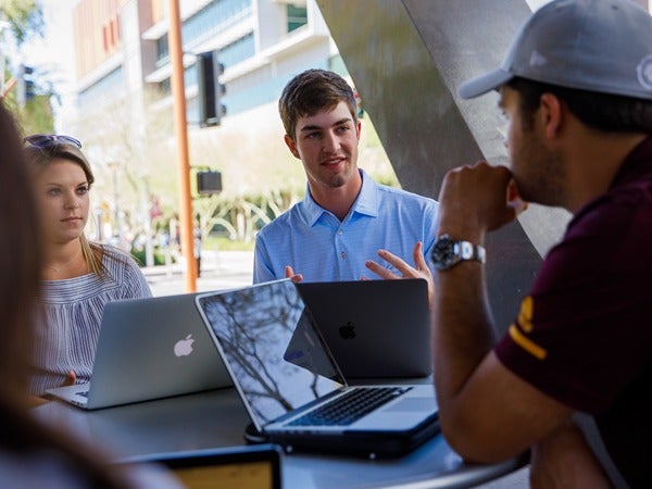 Three people having a discussion outdoors with laptops at a table, set on a sunny campus.