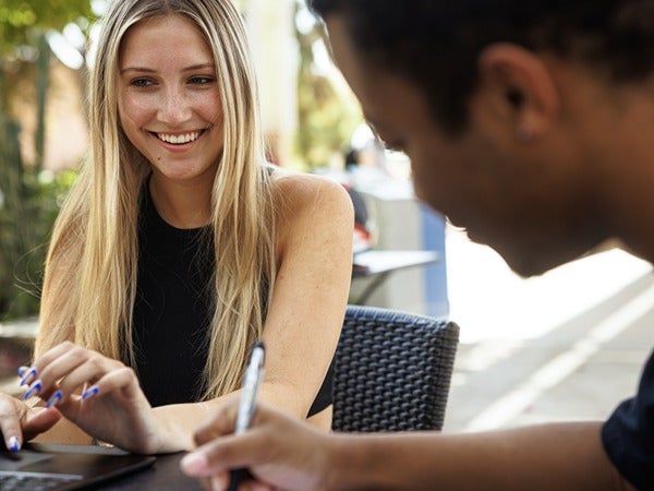 Two people sitting at a table outdoors, one smiling while using a laptop.