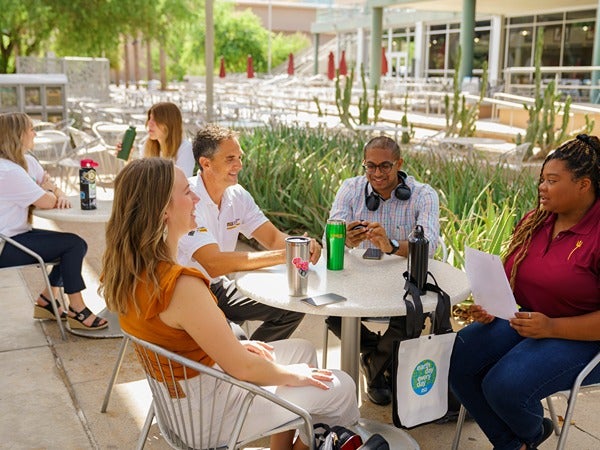 A group of five people seated at an outdoor table in conversation, surrounded by greenery and patio furniture.