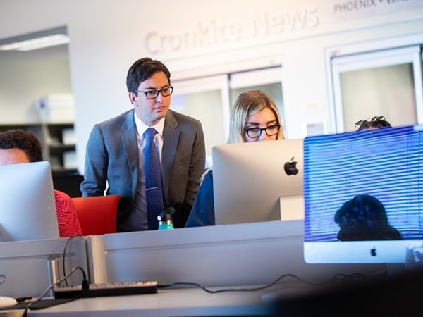 A man in a gray suit leans over to look at a computer screen with a seated woman in an office.