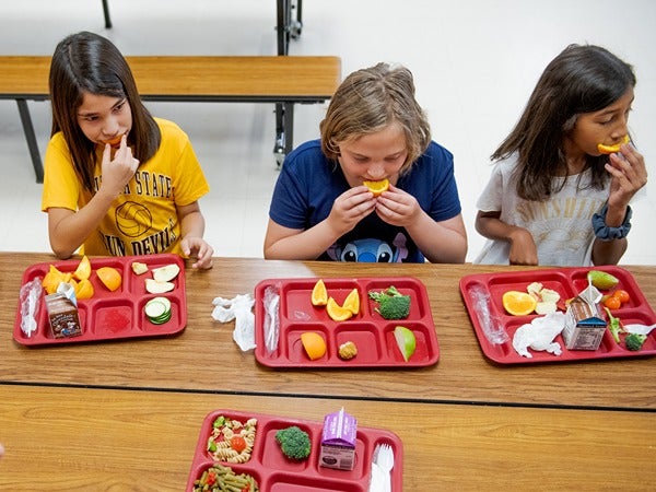 Three children eating lunch from red cafeteria trays with various fruits and vegetables.