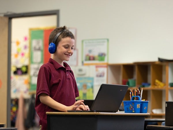 A boy with headphones smiling at a laptop in a classroom.