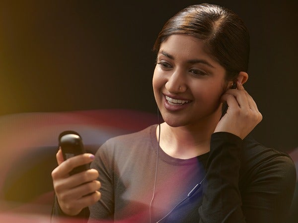 A smiling woman with earphones, holding a small device, against a dark, colorful background.