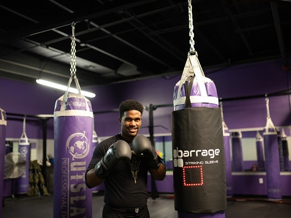 A person with boxing gloves poses by purple punching bags in a gym.