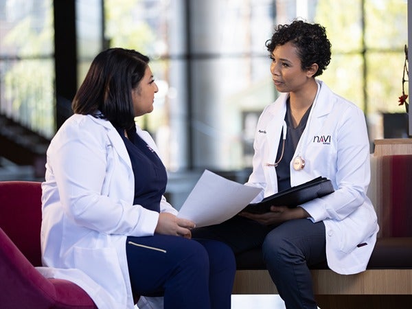 Two women in white lab coats converse while seated on a maroon couch in a brightly lit room.