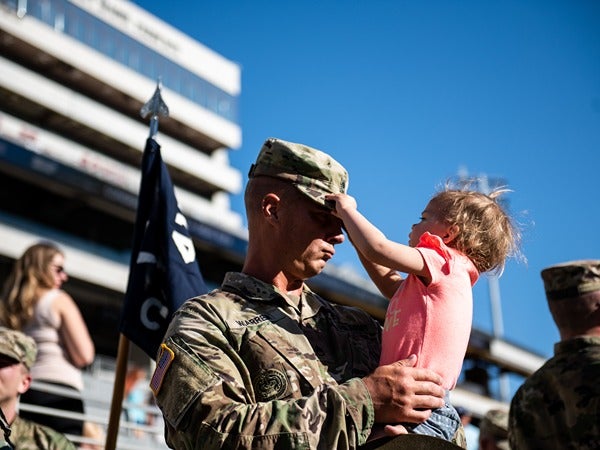 A soldier in camouflage holds a child who is touching his cap.