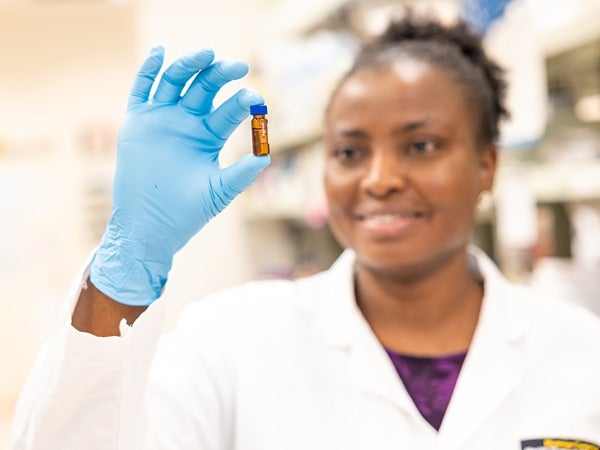 A person in a lab coat holding a small amber vial with a blue cap in a laboratory setting